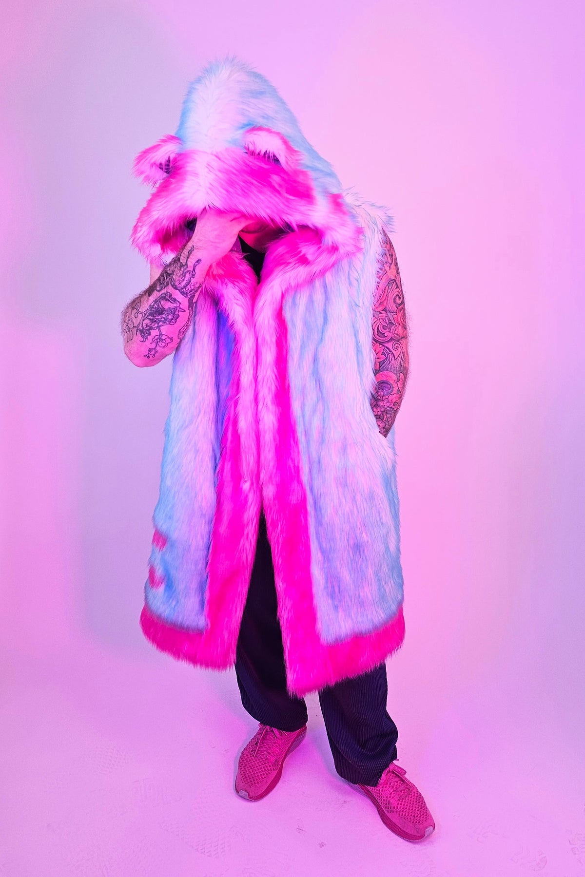 A person wearing a long hooded faux-fur vest with bright pink trim and a light blue body, standing against a pink-lit studio background. 