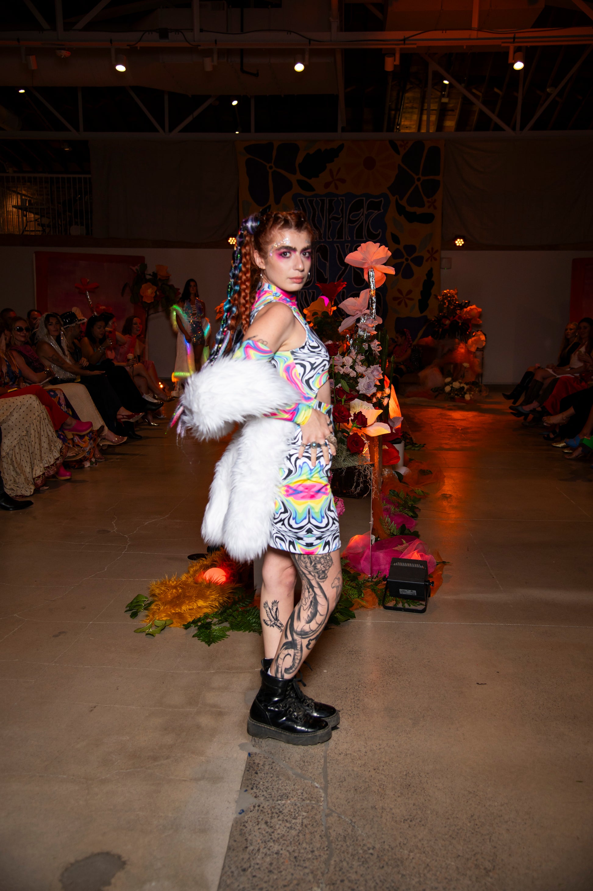 Model on a runway wearing a colorful outfit with a white fur accessory, surrounded by floral decorations.