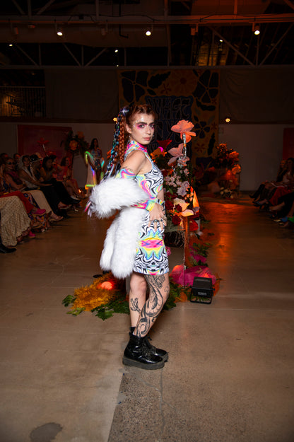 Model on a runway wearing a colorful outfit with a white fur accessory, surrounded by floral decorations.