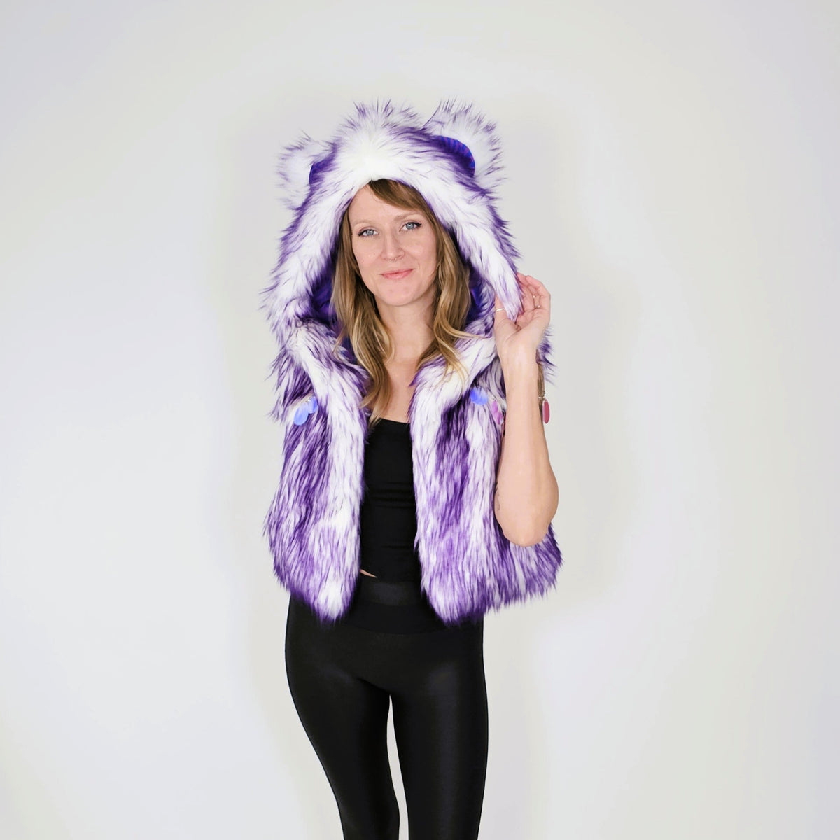 A woman modeling a purple and white faux fur hooded vest with bear ears, paired with a black top and black leggings, against a plain white studio background.
