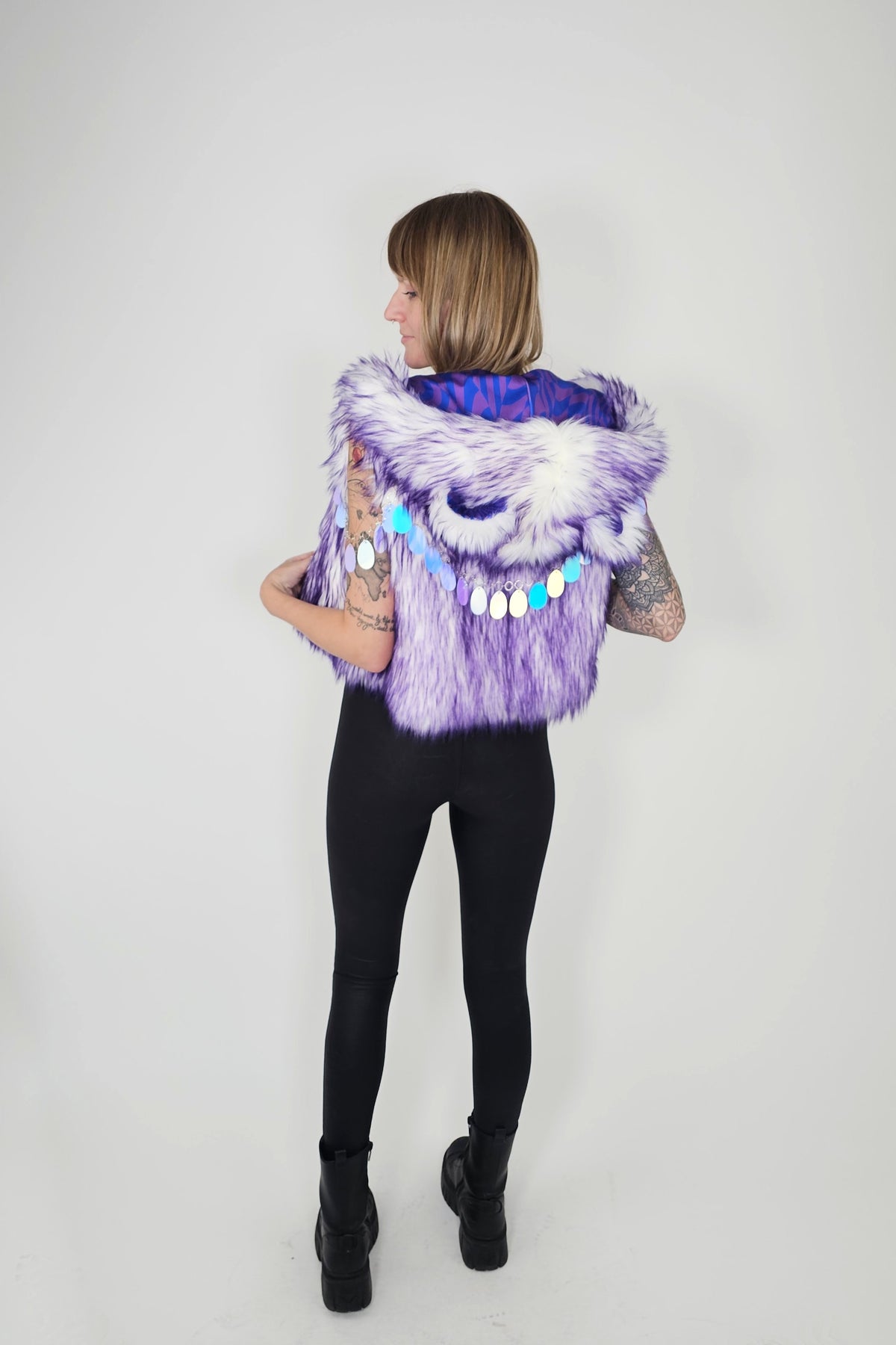 A woman shown from the back wearing a purple and white faux fur vest with a blue patterned lining and colorful bead accents, paired with black leggings and black boots, against a plain light gray background.