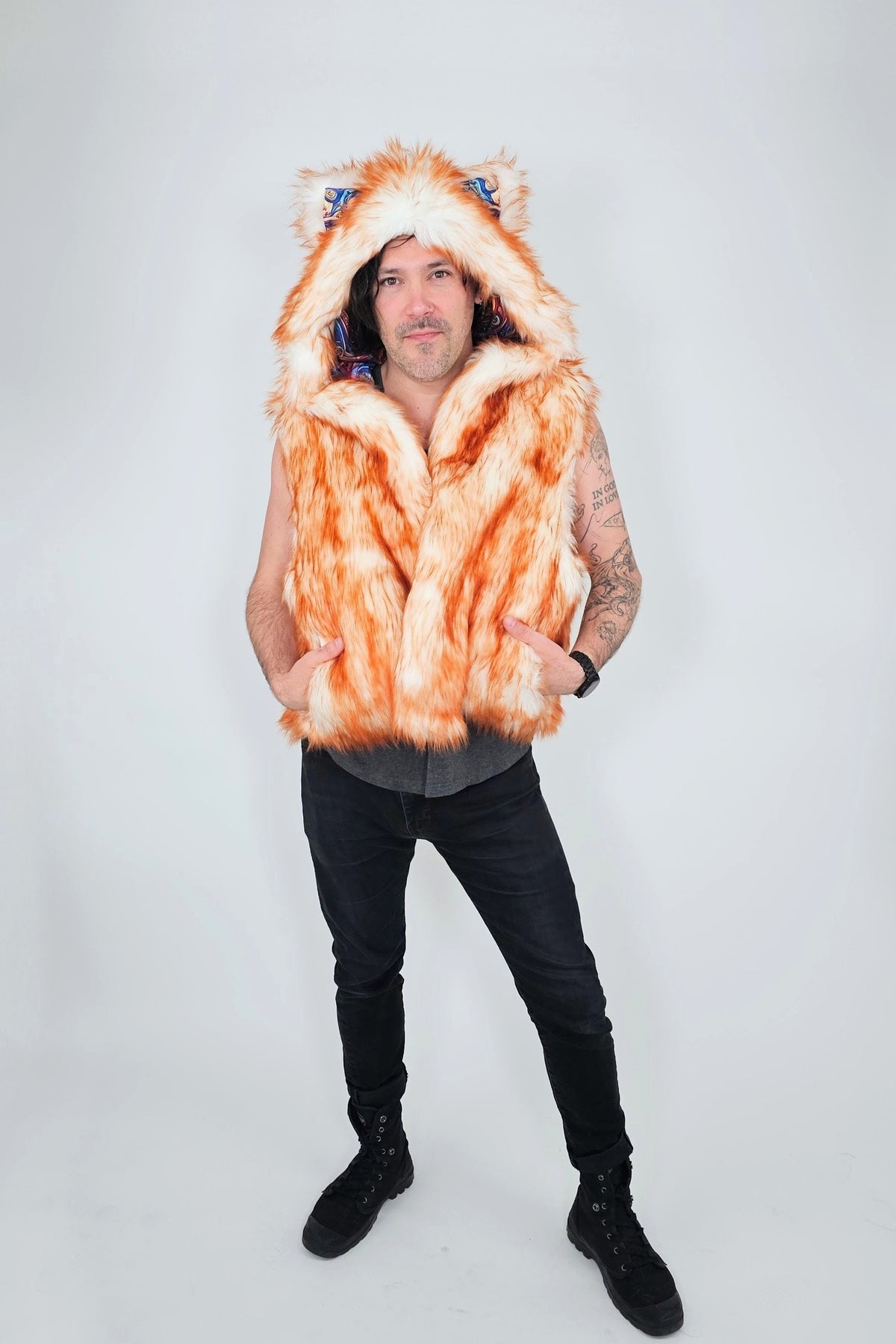 A man modeling a hooded orange and white faux fur vest with animal ears on the hood, paired with black pants and boots, against a light gray studio background.