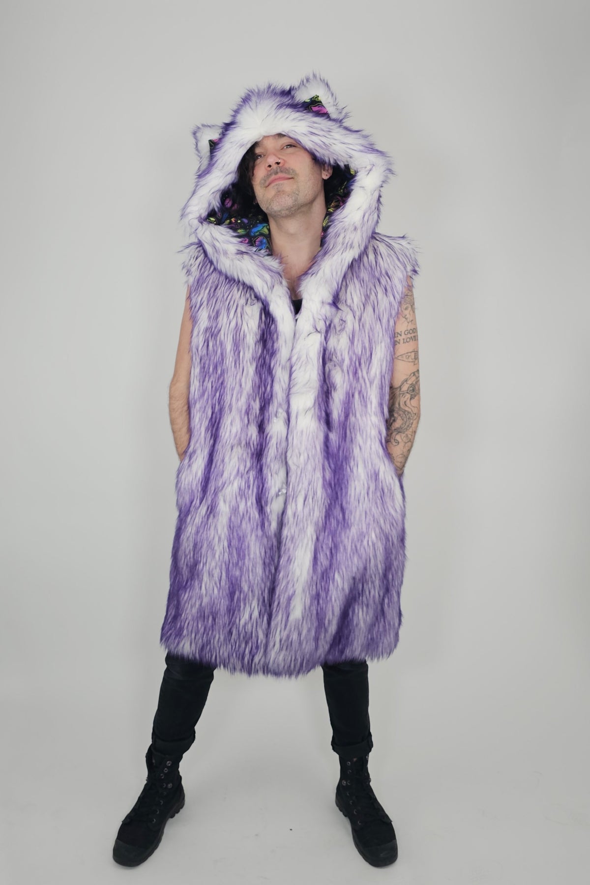 A person modeling a long purple and white faux fur hooded vest with cat ears on the hood, paired with black pants and black boots, against a light gray studio background.