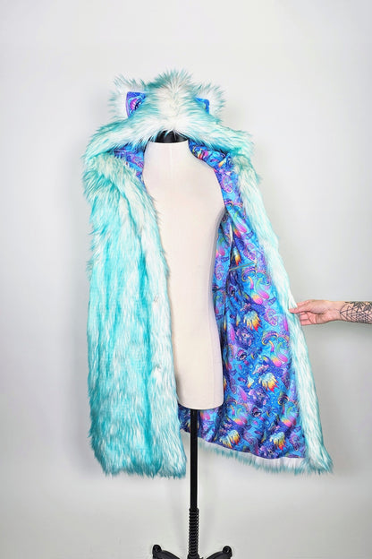 A hooded faux fur coat displayed on a cream dress form mannequin against a light gray background. The outer layer is light aqua/turquoise faux fur with cat-ear details, while the inner lining features a vibrant blue, purple, and pink abstract pattern.