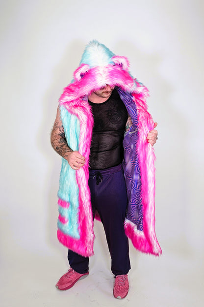 A person modeling a long hooded faux-fur vest with bright pink and light blue panels, a purple patterned lining, and fluffy trim, photographed against a plain light background.