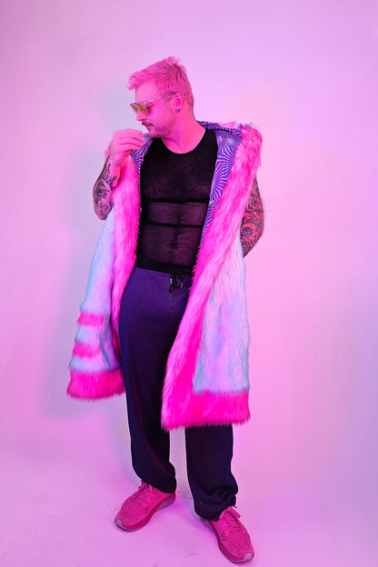 A person modeling a long, open-front faux fur coat with bright pink and light blue/white stripes, styled with a black mesh top, dark pants, and pink sneakers against a pink-lit studio background.