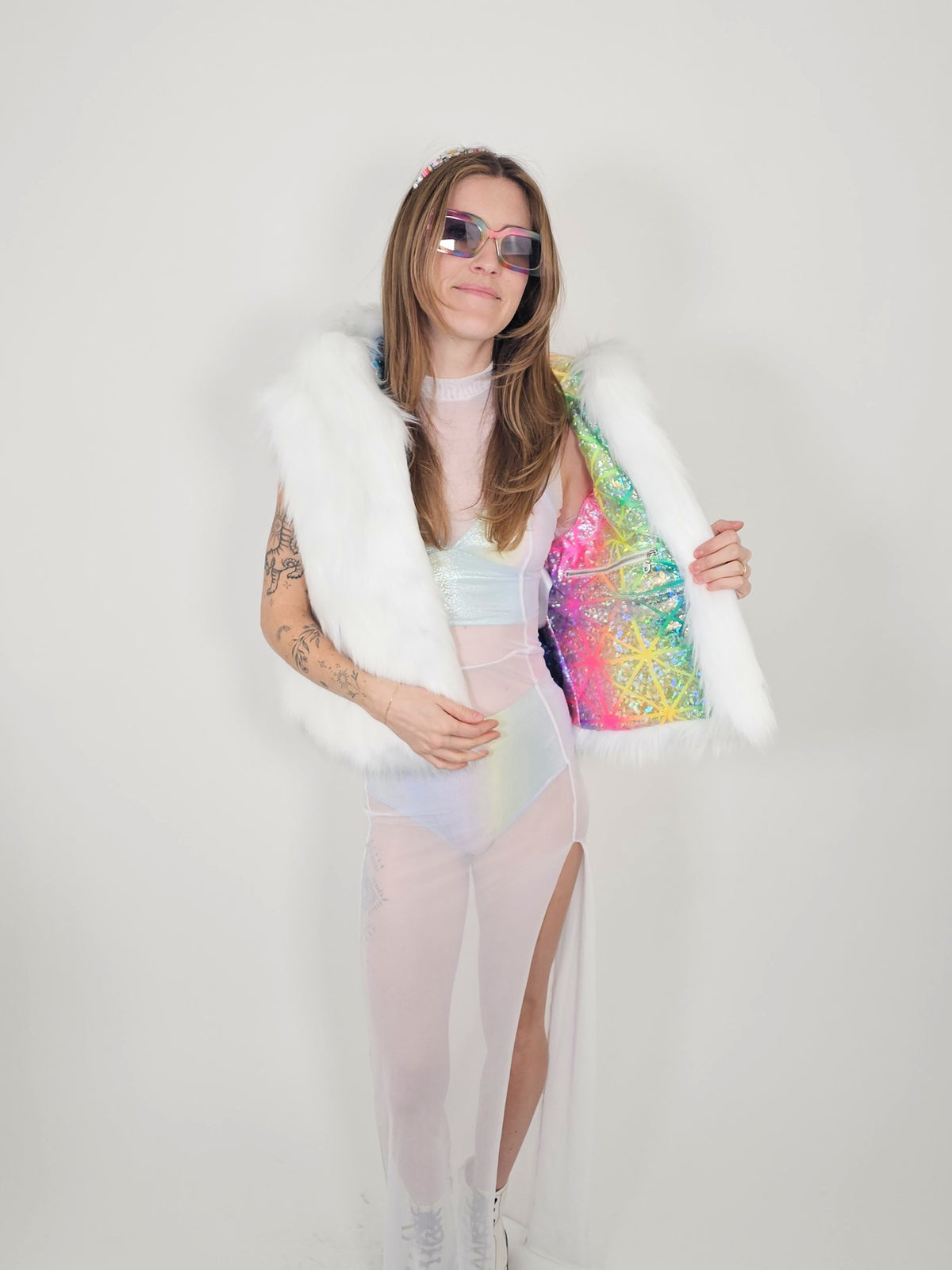 A woman modeling a white faux fur vest over a pastel iridescent bodysuit, holding open a bright iridescent pink, yellow, and blue sequined jacket. She is wearing pink-tinted sunglasses and standing against a plain light gray background.