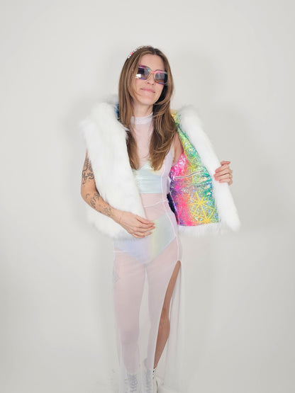 A woman modeling a white faux fur vest over a pastel iridescent bodysuit, holding open a bright iridescent pink, yellow, and blue sequined jacket. She is wearing pink-tinted sunglasses and standing against a plain light gray background.