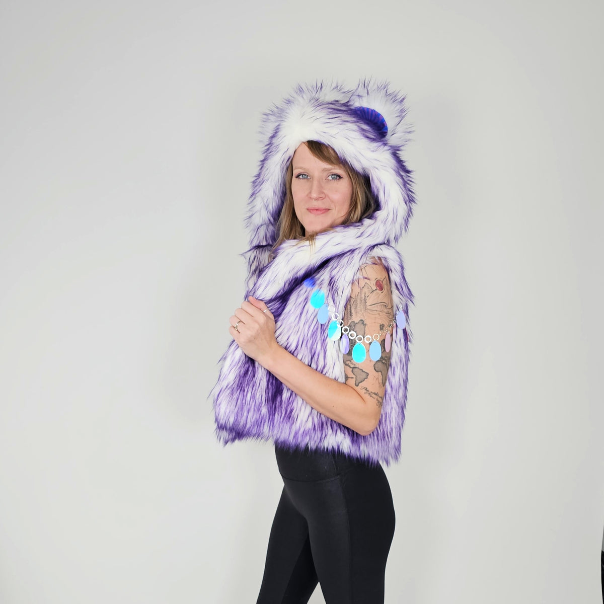 A woman modeling a purple and white faux fur hooded vest with a pom-pom on the hood, paired with black leggings, against a plain light gray background.