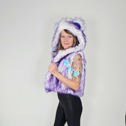 A woman modeling a purple and white faux fur hooded vest with a pom-pom on the hood, paired with black leggings, against a plain light gray background.